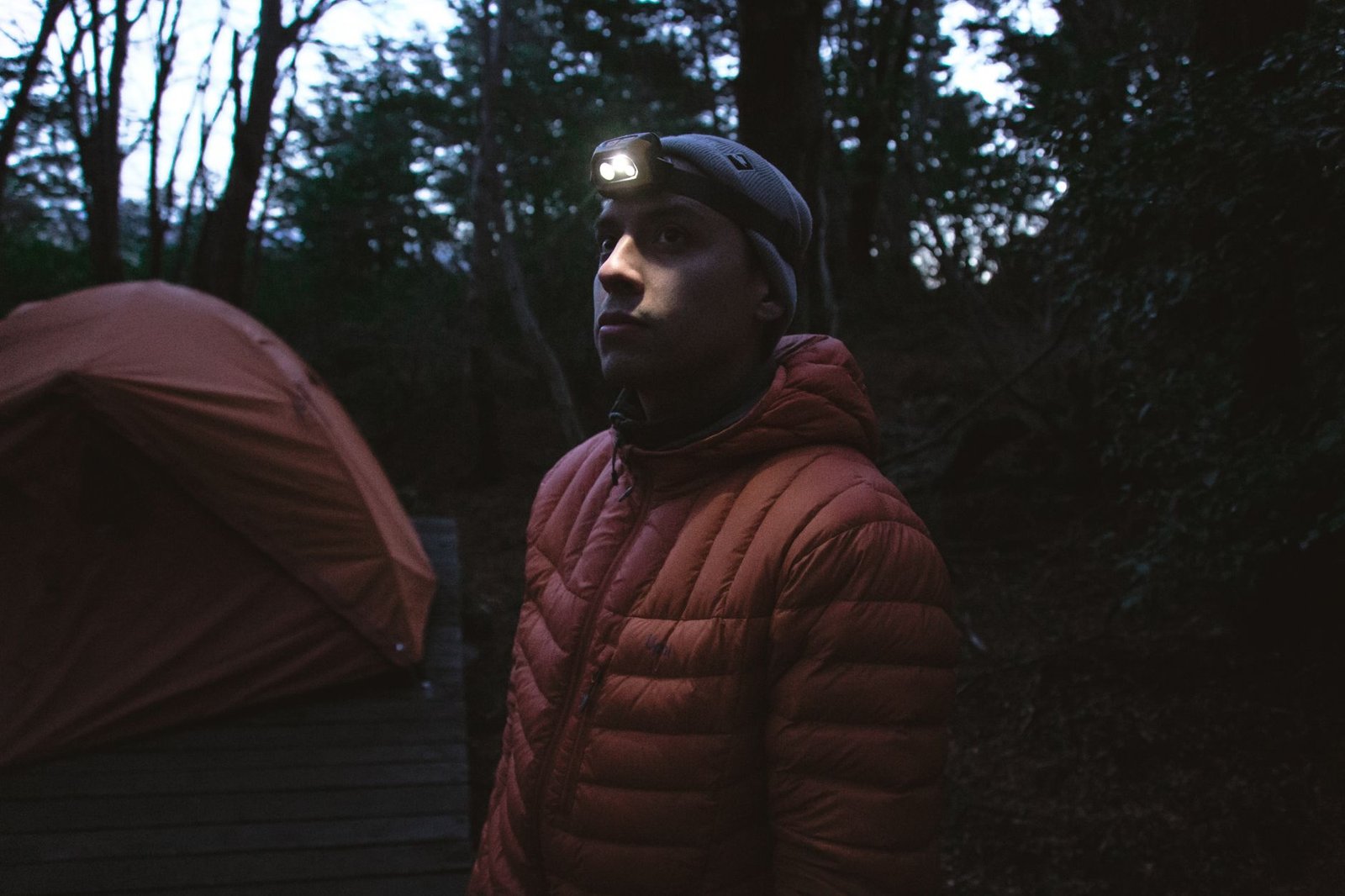 Man met een brandende hoofdlamp naast een tent in het bos bij schemering