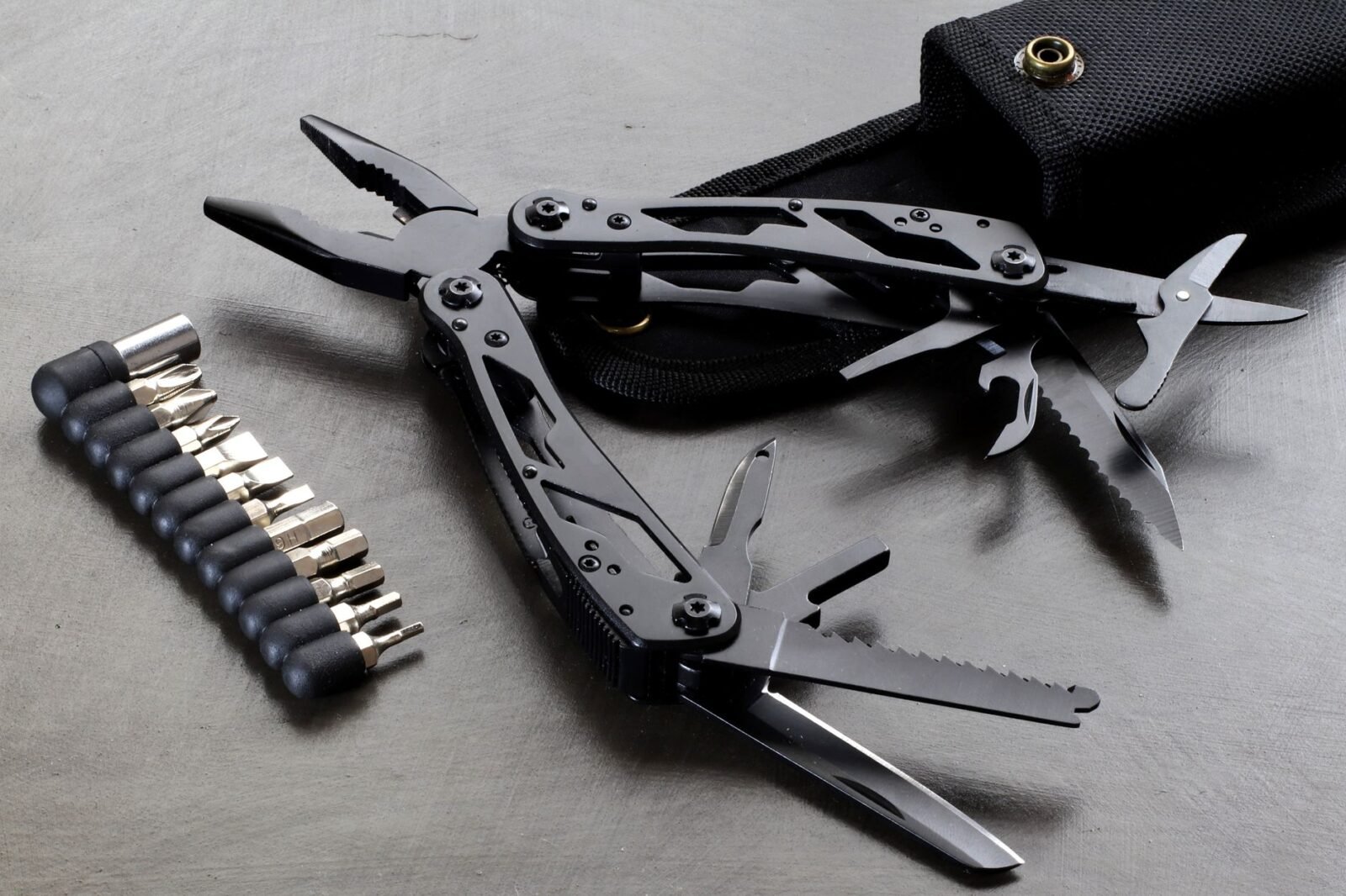 Zwarte multitool met uitgeklapte tang, zaag en mesjes naast een set bits